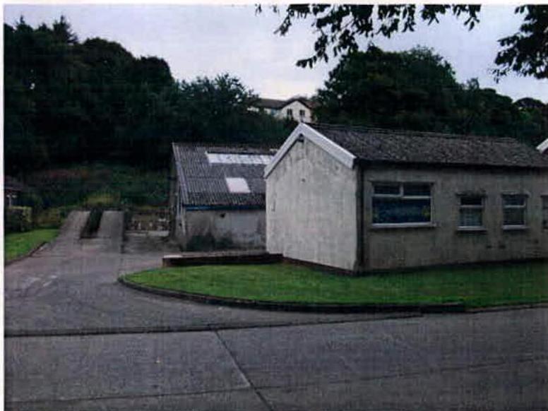 Exterior photograph showing a single-story building with a pitched roof and white rendering, situated next to a paved driveway with trees in the background.