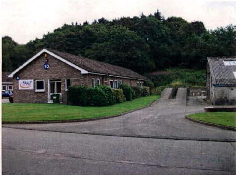 A photograph showing a single-story brick building with a 'RALLY' sign and a smaller adjacent outbuilding, set against a wooded hillside with a driveway in the foreground.