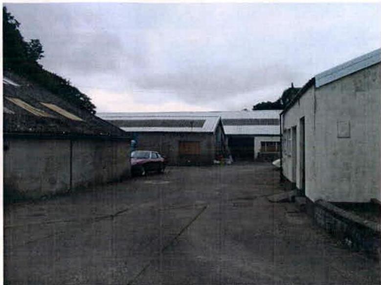 This is a photograph showing a paved yard area surrounded by several buildings, including a long white structure in the background and smaller structures on the sides.