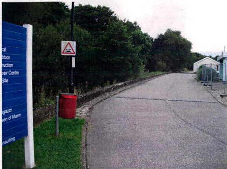 A photograph showing a paved access road leading to a site, featuring a blue sign on the left, a speed bump warning sign, and white buildings in the distance.