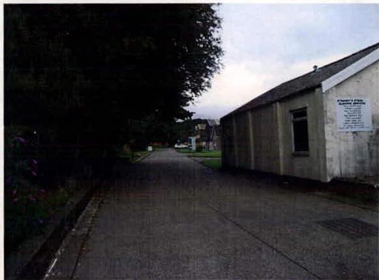 A photograph showing a paved access road alongside a long, single-story industrial building with a sign on its exterior wall.
