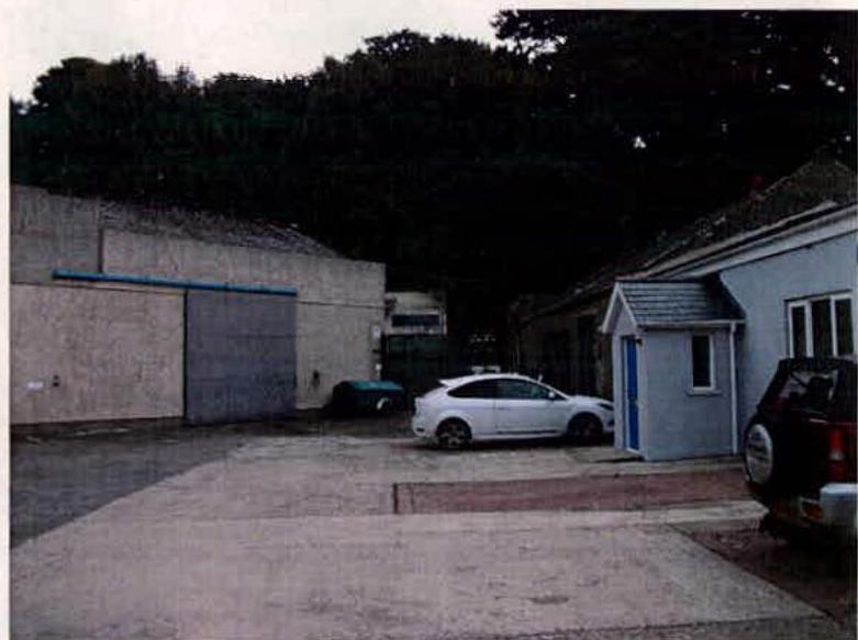 A photograph showing an industrial yard with a large concrete building featuring a roller shutter door and a smaller white structure with parked cars.