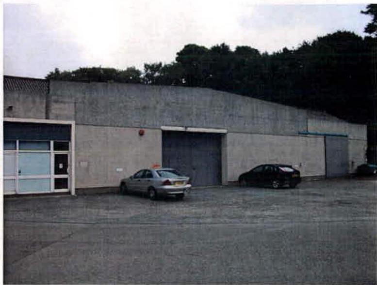 A photograph showing a large, grey concrete industrial building with roller shutter doors and a pedestrian entrance, with cars parked on the paved foreground.