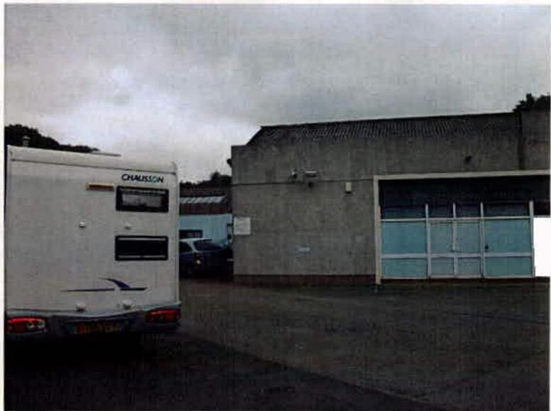 A photograph showing a white caravan parked on a paved area next to a grey industrial building with a large roller shutter door.