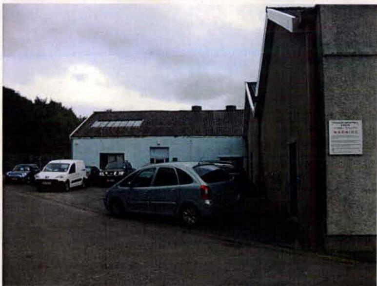 A street-level photograph showing industrial buildings with cars parked in the foreground, representing the site of the proposed demolition.