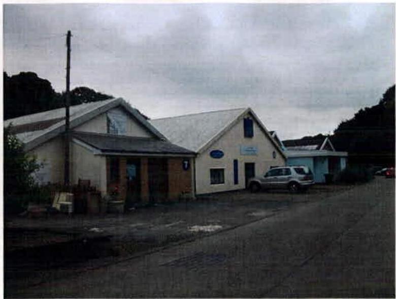 A street-level photograph showing a row of white industrial or commercial buildings with pitched roofs. A silver car is parked in front on a paved area.