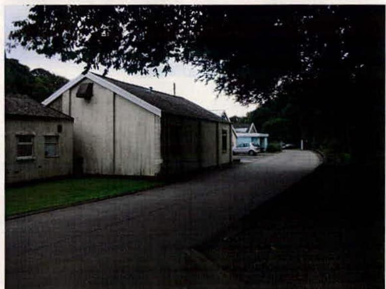 Exterior photograph showing a white industrial-style building and adjacent structures along a paved road, likely the buildings proposed for demolition.