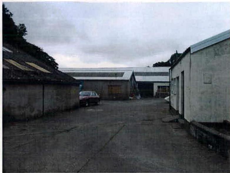 A photograph showing a paved industrial yard surrounded by several buildings, including a long white structure in the background and older structures on the sides.