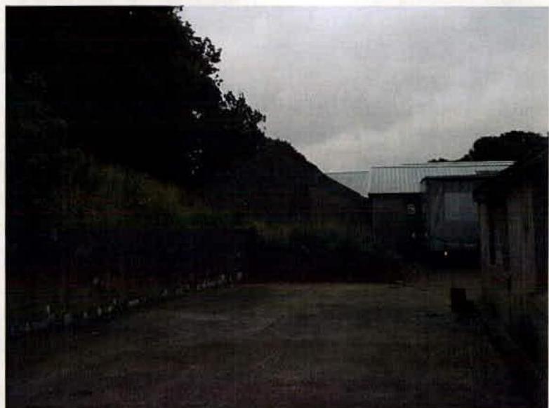 A grainy photograph showing an industrial site with a paved yard, a grassy slope on the left, and buildings with corrugated roofs in the background.