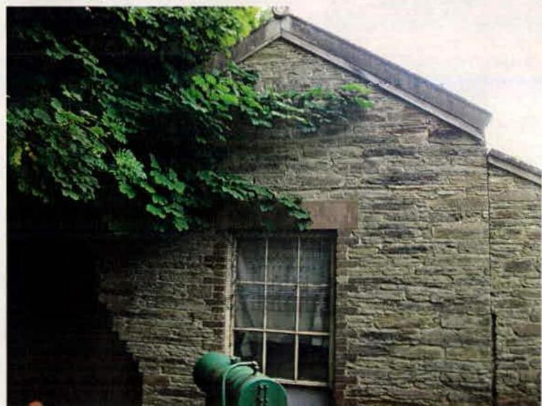 A photograph showing the exterior stone wall of an existing building with a multi-paned window and overhanging vegetation, likely the laundry building proposed for demolition.