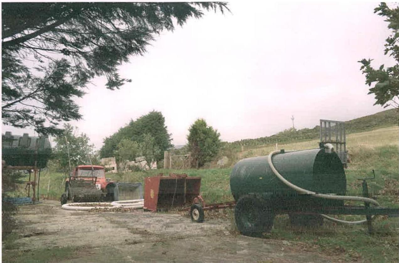A photograph depicting agricultural equipment, including a red tractor and a large green tanker, situated on a dirt track in a rural landscape.