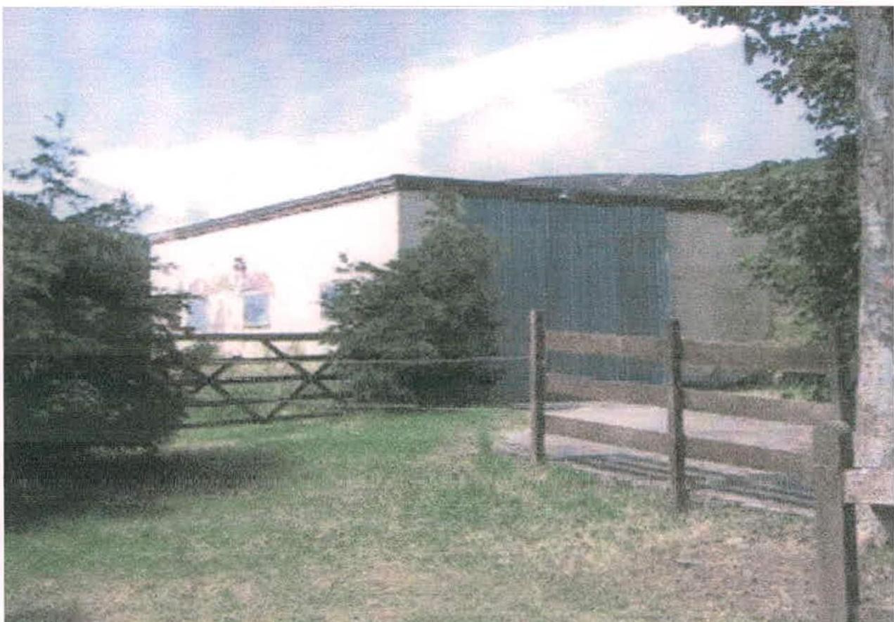 A photograph showing a large agricultural building or barn with mixed cladding, situated behind a wooden fence in a rural setting.