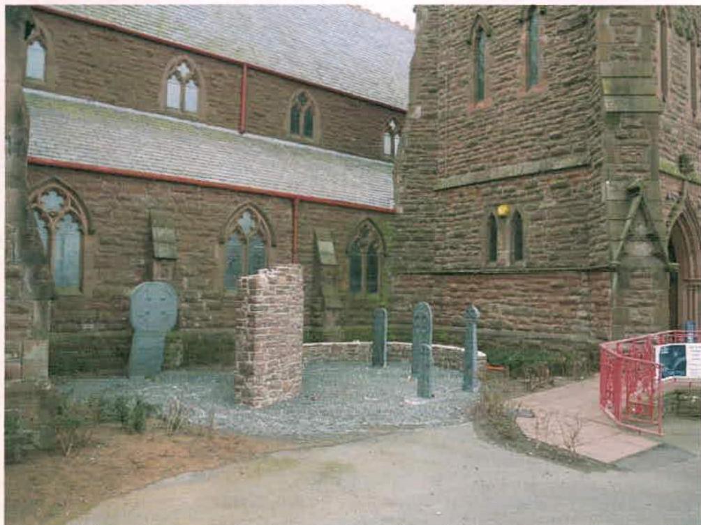 A photograph showing the exterior of a historic stone building, likely a church, with a graveyard area containing headstones and a low stone wall.