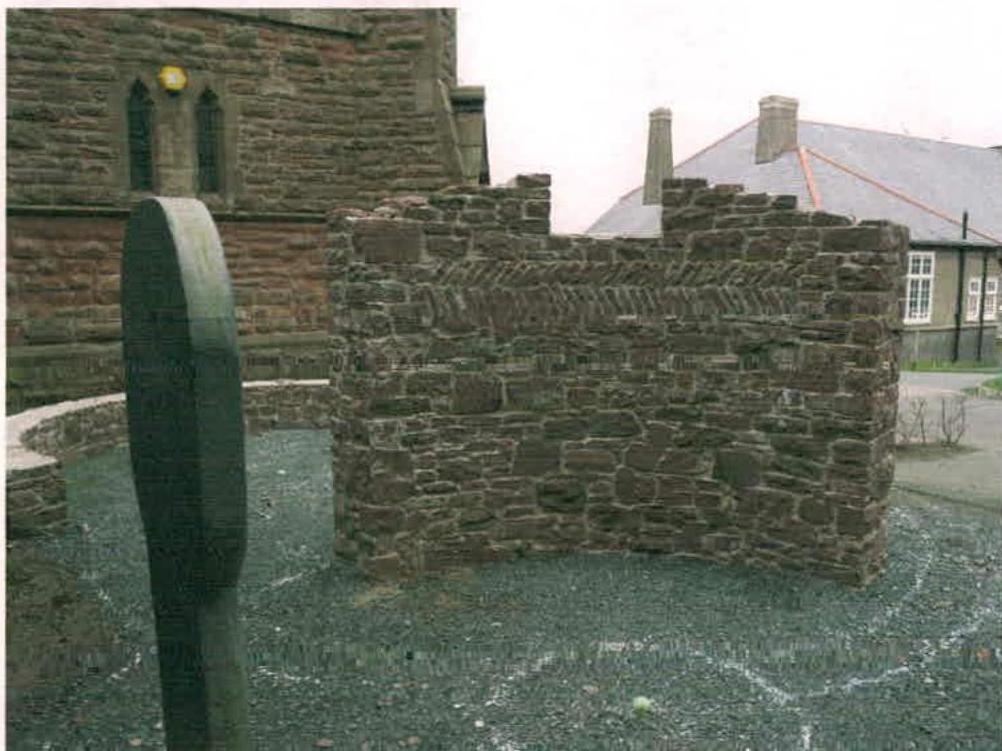 A photograph showing a low stone wall and gravel ground in a garden area, with a stone building and a house in the background.