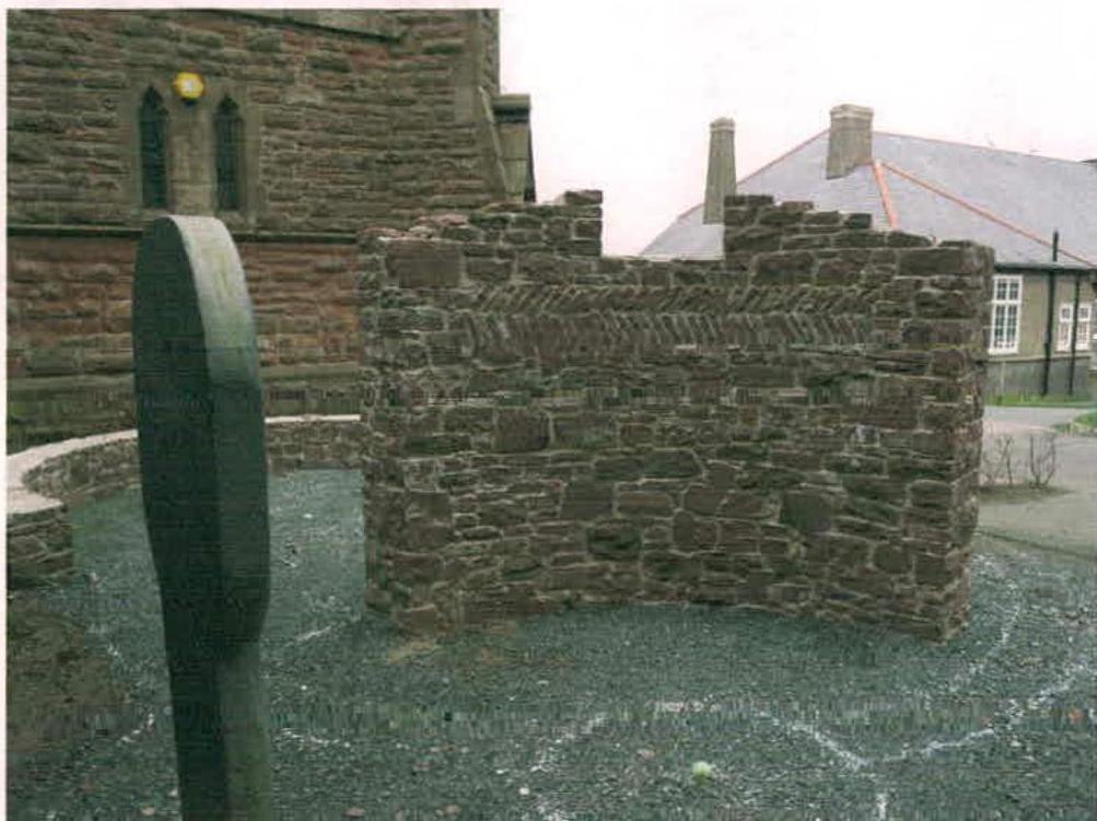 A photograph showing a low stone wall or boundary feature in a gravel area, with a historic stone building on the left and a residential house in the background.
