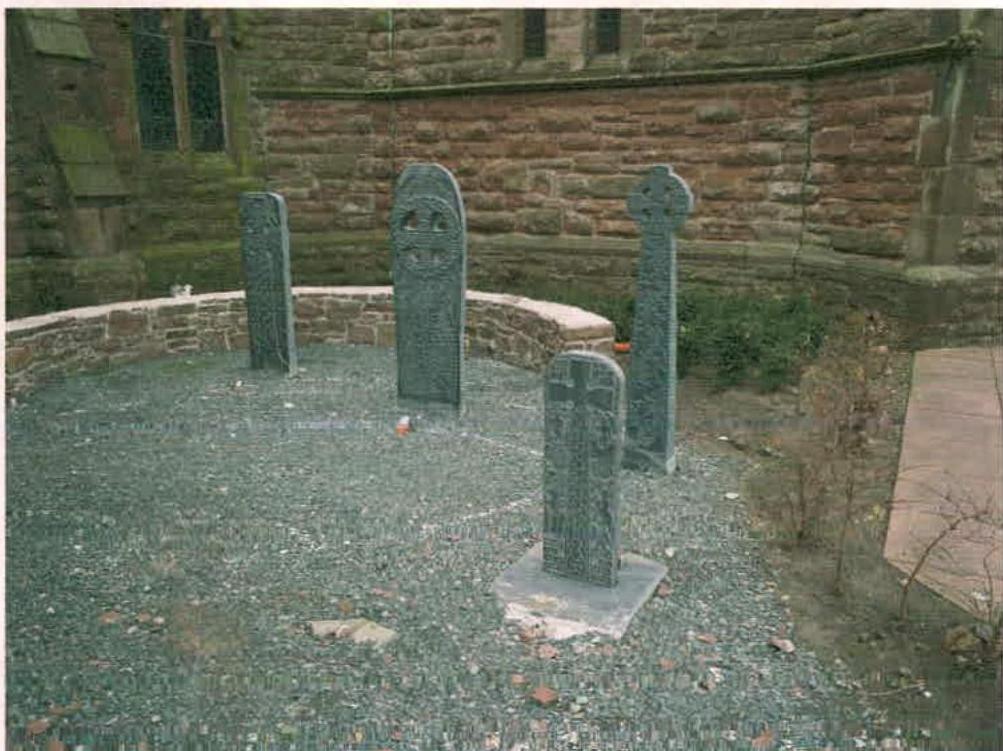 A photograph showing a gravel garden area containing four upright stone slabs with Celtic cross carvings, set against a historic stone wall.