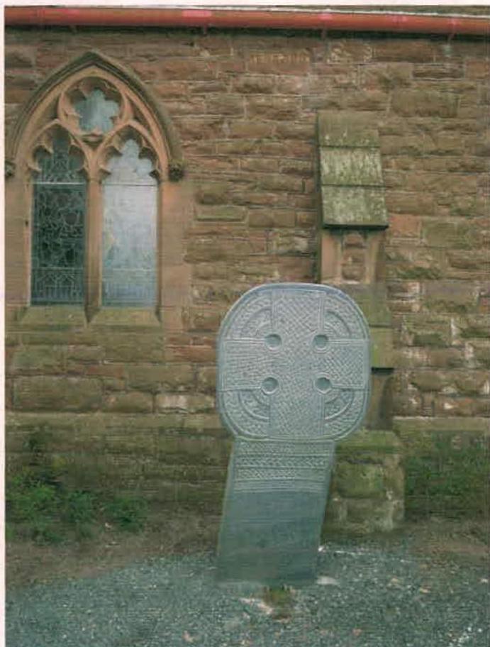 A photograph showing a stone Celtic cross standing in front of a stone building with a Gothic-style window.