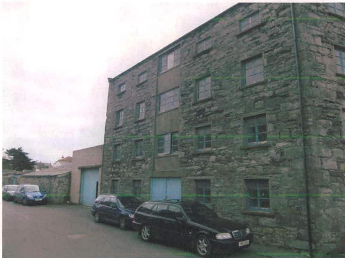 A photograph of a large, multi-story stone warehouse building with cars parked in front and a smaller outbuilding to the left.