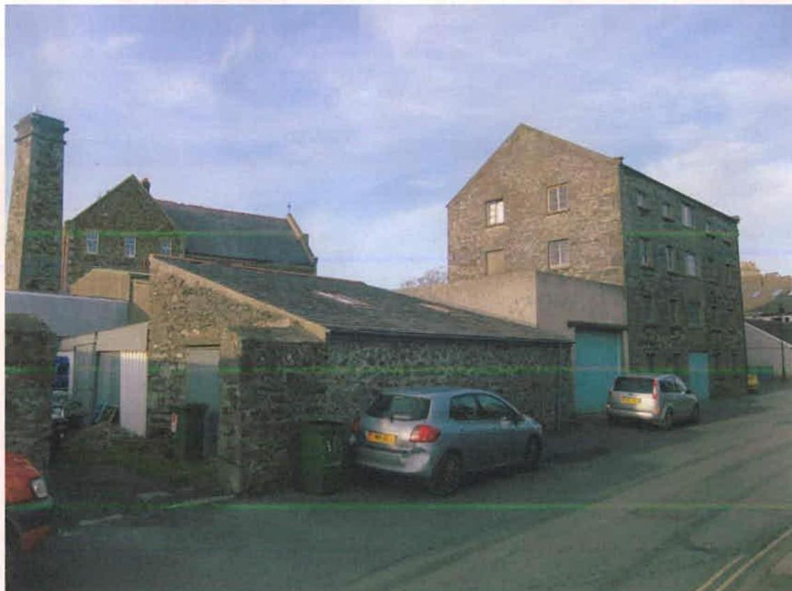 This photograph shows the existing site, featuring stone industrial buildings including a tall warehouse and a lower workshop structure with vehicles parked nearby.