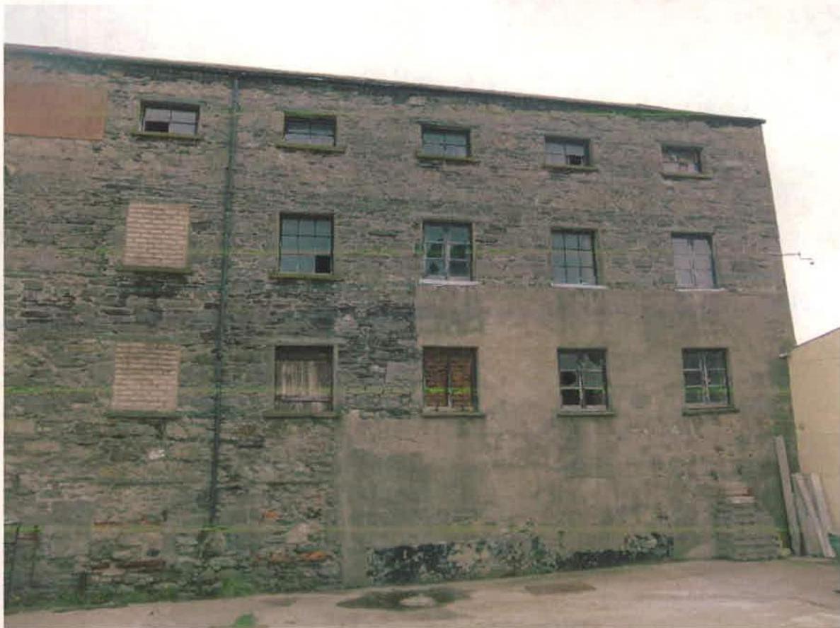 A photograph showing the exterior facade of a large, multi-story stone warehouse building with mixed masonry and boarded windows.