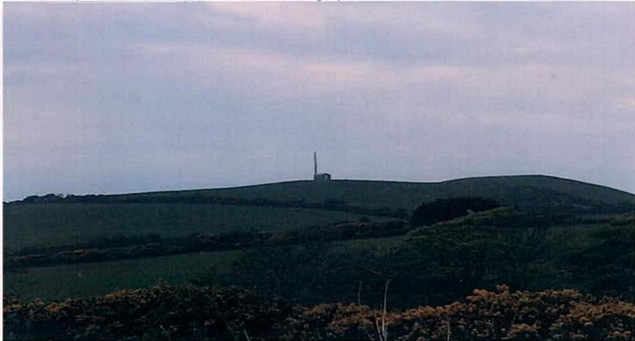 A landscape photograph showing a grassy hill with a telecommunications mast and small equipment building on the ridge against an overcast sky.