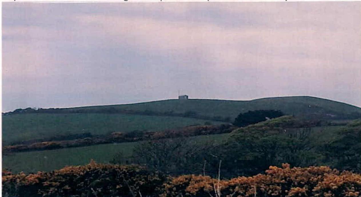 A photograph of a rural landscape showing a green rolling hill with a small structure visible on the ridge against an overcast sky.