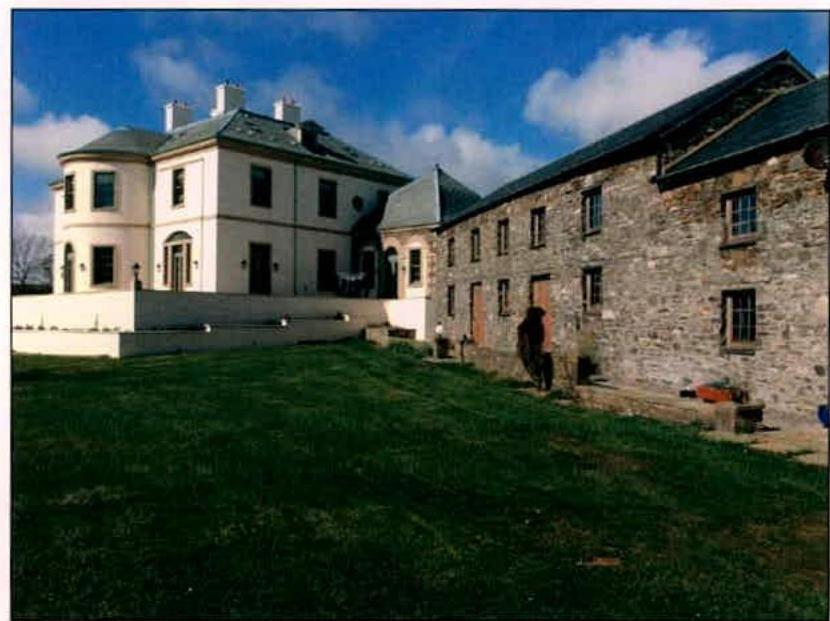 A photograph showing a large white detached house adjacent to a long stone outbuilding or barn, set against a blue sky with a grassy lawn in the foreground.