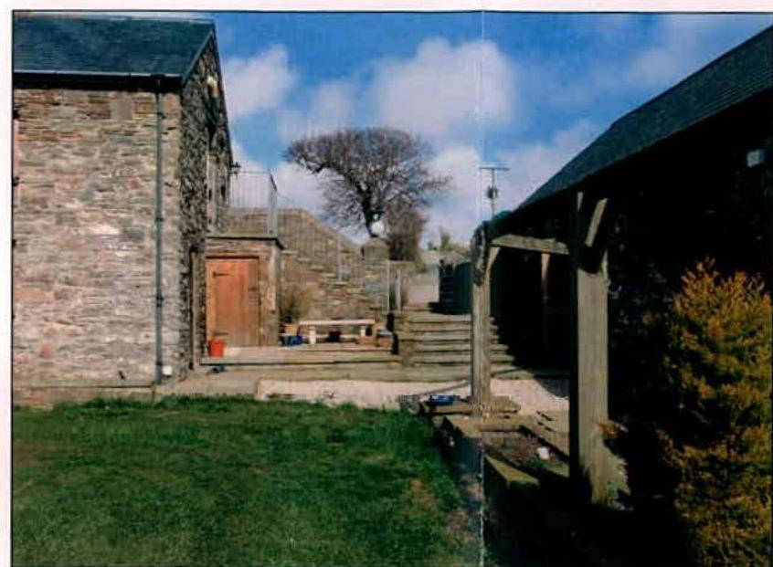 A photograph showing the rear garden area of a property featuring a stone building on the left and a wooden structure on the right.