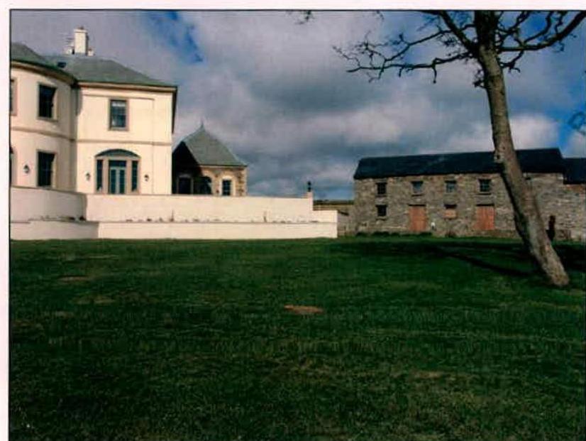 A photograph showing a large white detached house on the left and a long stone barn-like outbuilding on the right, separated by a smaller structure, all set behind a large grassy lawn.