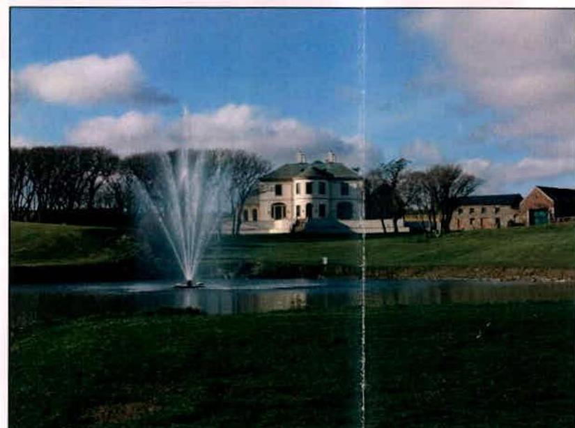 A photograph showing a large white detached house situated behind a pond with a fountain. The image captures the rural setting with outbuildings visible to the right.