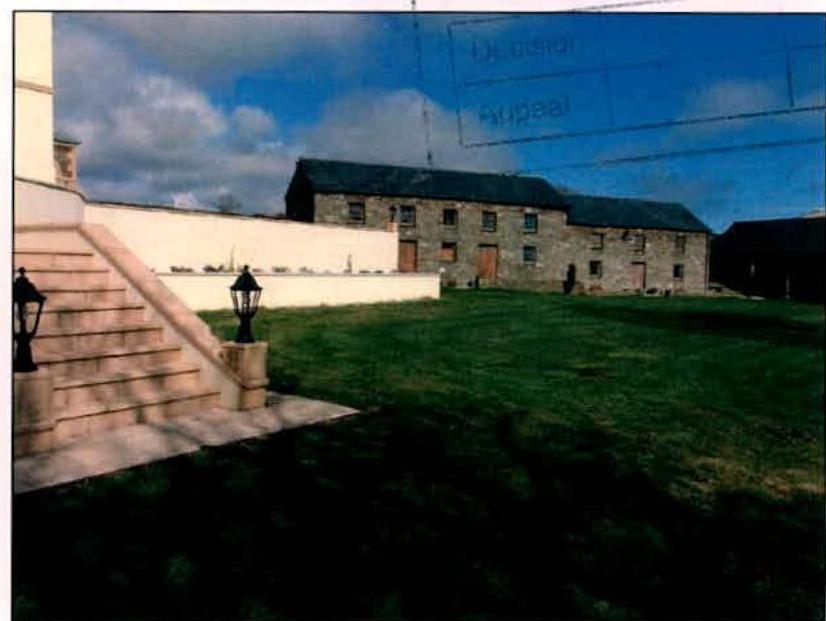 A photograph showing a large stone barn-like building in the background with a grassy lawn in the foreground and stone steps leading up to a white wall on the left.