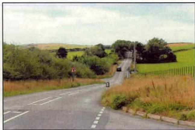 A photograph showing a rural road curving uphill through green fields and trees, with a car driving away in the distance.