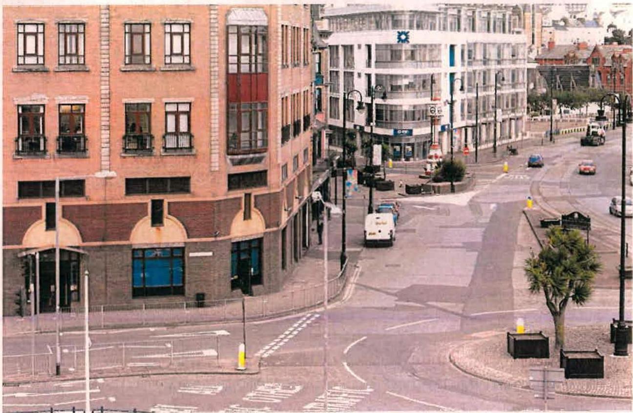 A street-level photograph showing a town center scene with multi-story commercial buildings, a paved road with traffic, and street furniture, likely depicting the Douglas Promenade area.