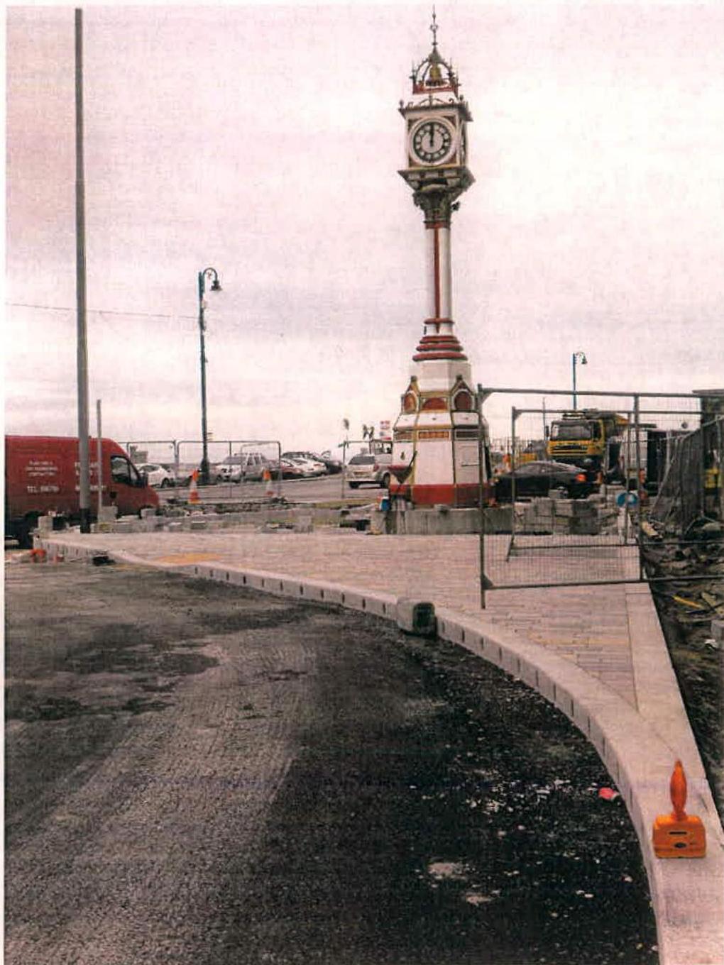 A photograph showing construction work on a coastal promenade featuring a prominent clock tower. New paving and curbs are being installed around the base of the tower with construction vehicles nearby.