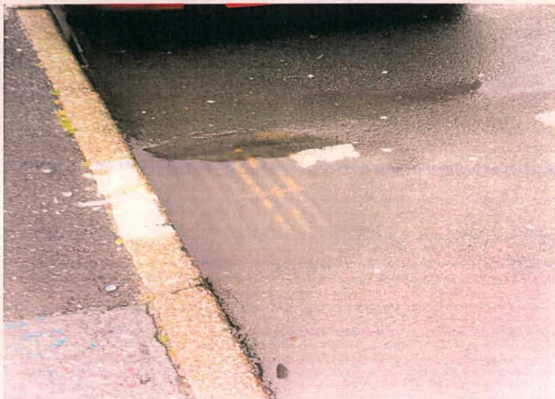 A close-up photograph of a wet asphalt road surface showing double yellow lines and a curb.