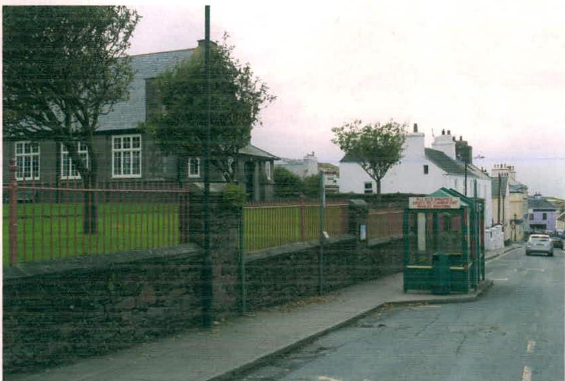 A street-level photograph showing a stone wall and fence bordering a grassy area with a large building, likely the cathedral grounds, and an existing green bus shelter on the sidewalk.