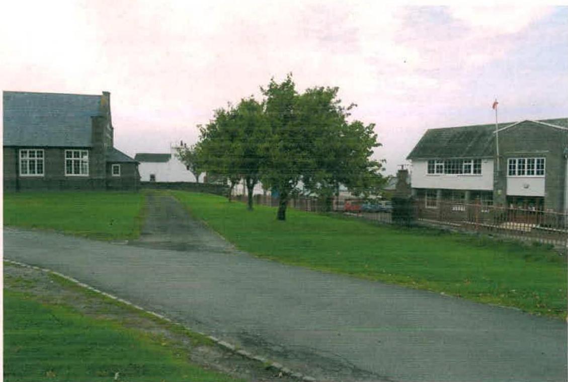 A photograph showing a road scene with a stone building on the left and a white building on the right, separated by a grassy area with trees and a path leading towards the stone building.