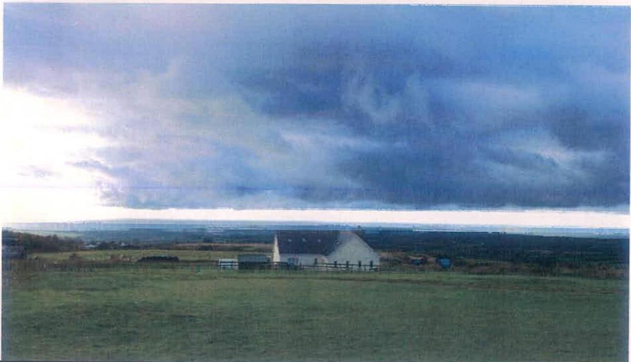 A photograph showing a rural landscape with a white building in the middle ground and a view of the sea in the distance under a cloudy sky.