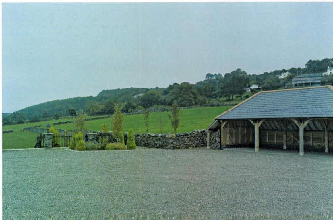 A photograph showing a large wooden outbuilding with a slate roof situated next to a gravel driveway in a rural setting.