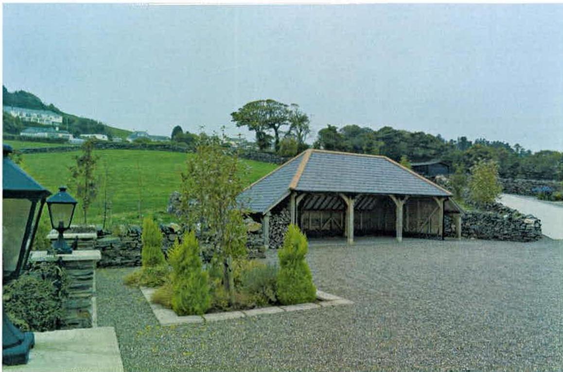 A photograph showing a large, open-sided detached garage with a slate roof situated on a gravel driveway in a rural setting.