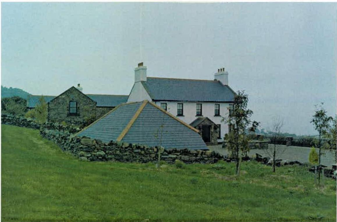 A photograph of a rural property featuring a white two-story house, a stone outbuilding, and a structure with a pyramid-shaped slate roof in the foreground.