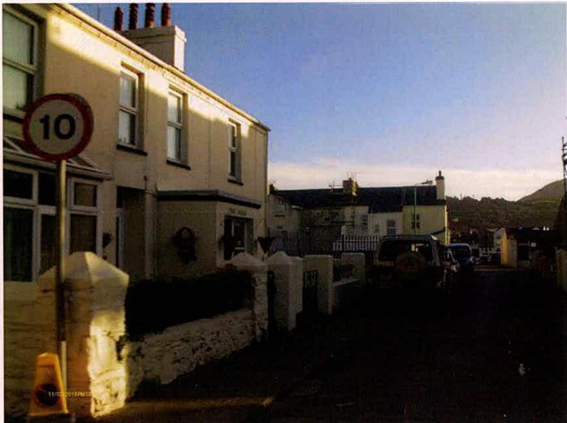 A street-level photograph showing existing buildings, including a two-story structure with a speed limit sign, and parked vehicles along a road.