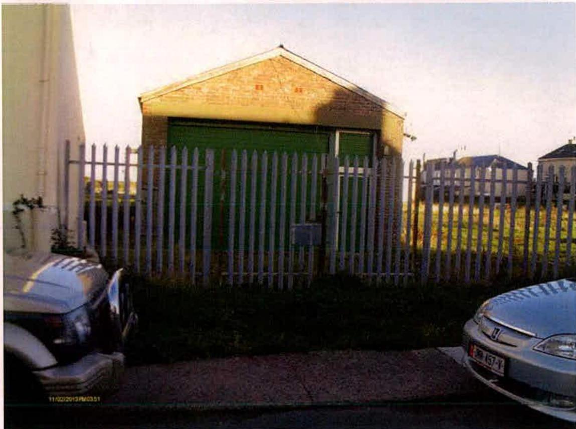 A photograph of a single-story brick building with a green roller shutter door, enclosed by a white picket fence with cars parked in the foreground.