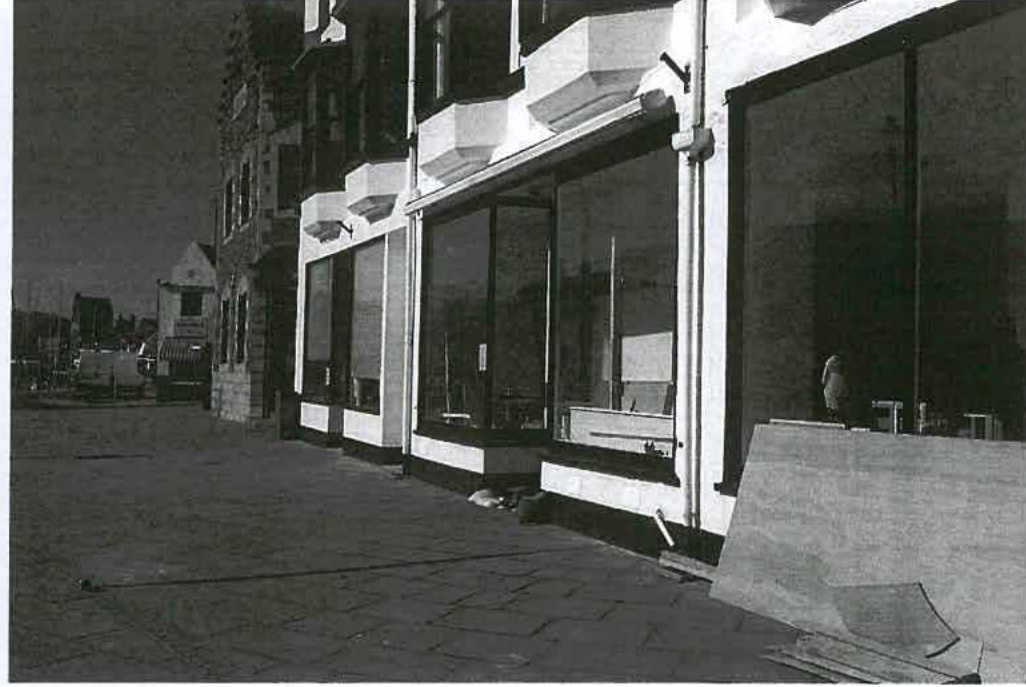 A black and white photograph showing the exterior facade of a terrace of commercial units with large shop windows and a paved forecourt. The background suggests a coastal location with masts visible near a harbor.