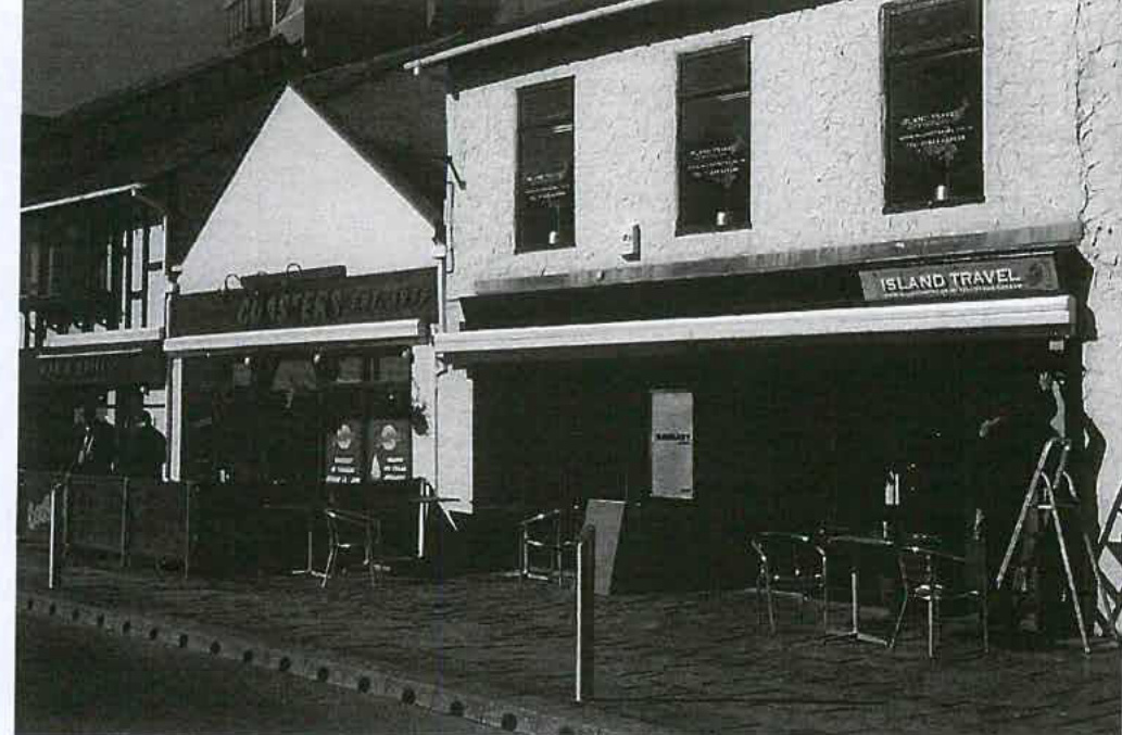 A black and white photograph showing the street-level view of commercial properties with existing outdoor seating furniture.