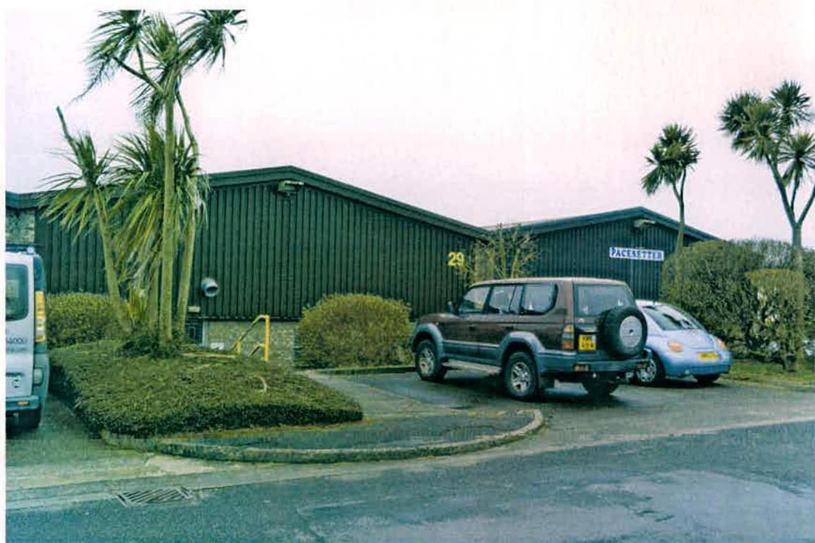 A street-level photograph showing the exterior of a dark green industrial unit with corrugated cladding and palm trees. Parked vehicles are visible in the paved area in front of the building.