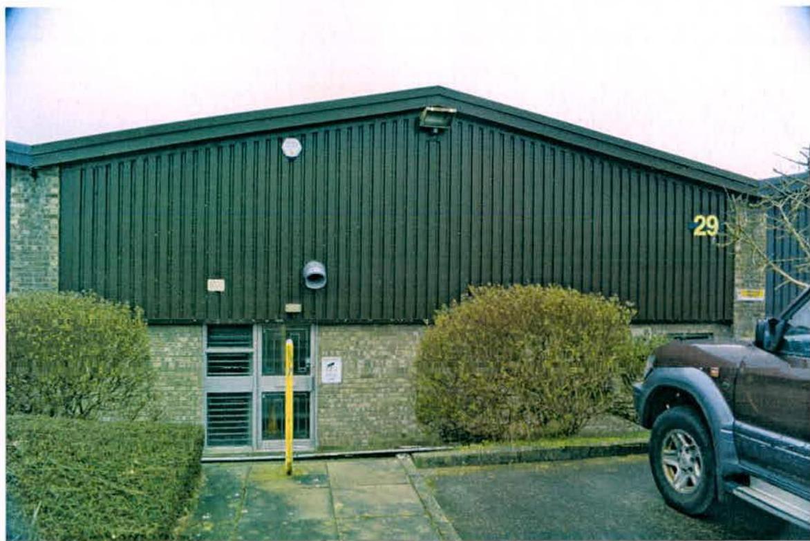 Exterior photograph of a single-story industrial unit featuring dark green cladding, brickwork, and a main entrance with a parked vehicle nearby.