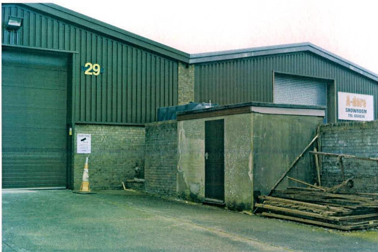 A photograph showing the exterior of a green industrial unit with corrugated metal cladding and a large roller shutter door, alongside a smaller attached structure and paved forecourt.