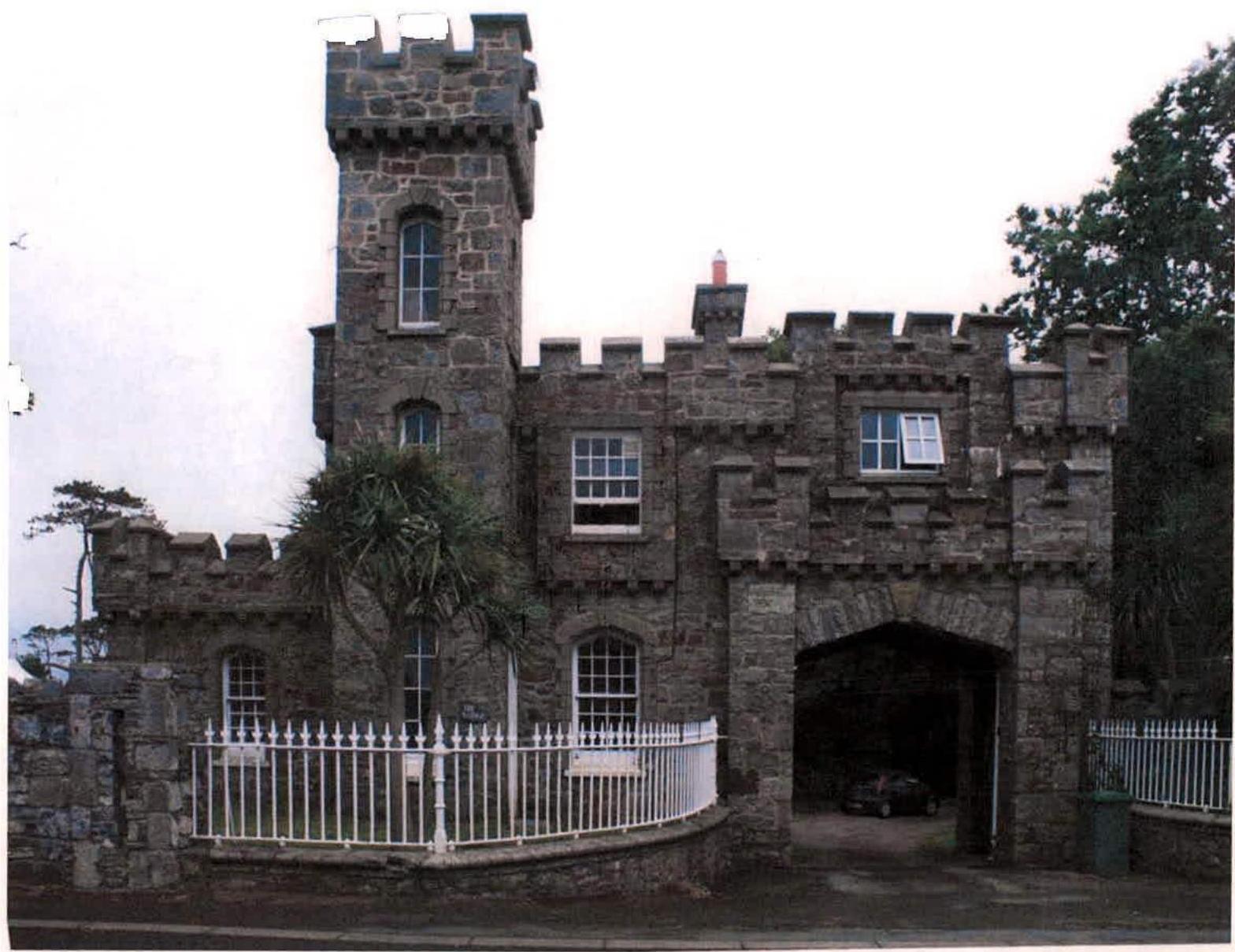 A photograph of a distinctive stone building resembling a small castle or gatehouse with crenellations, a tower, and a large arched entrance.