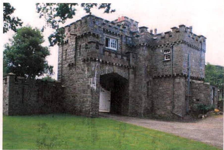 A photograph of a large stone gatehouse structure featuring castle-like crenellations and a central arched driveway entrance.